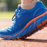 Gros plan sur une chaussure de running bleue et orange Asics GT-2000 13 en plein mouvement sur une piste d'athlétisme.
