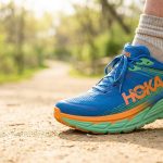 Une chaussure de running Hoka Gaviota 5 bleue et verte portée sur un sentier en terre en extérieur.