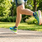 Un coureur porte des chaussures de running bleues et jaunes en pleine foulée sur un chemin de parc ensoleillé.
